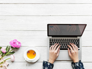 Hands typing on a laptop on a white wooden table, accompanied by a cup of tea and pink flowers. The screen shows an email inbox representing when to use email in business.