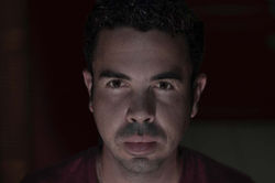 Face 6. Horizontal color close-up of a man looking directly at the camera, with low-key indoor night