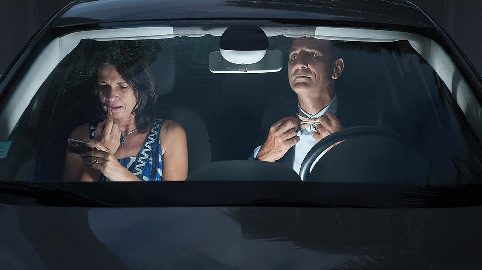 An elegantly dressed man adjusting his bow tie and a woman applying lipstick, sitting in a car.