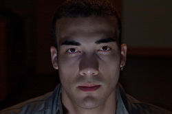 Face 1. Horizontal color close-up of a man looking directly at the camera, with low-key indoor night