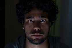 Face 2. Horizontal color close-up of a man looking directly at the camera, with low-key indoor night