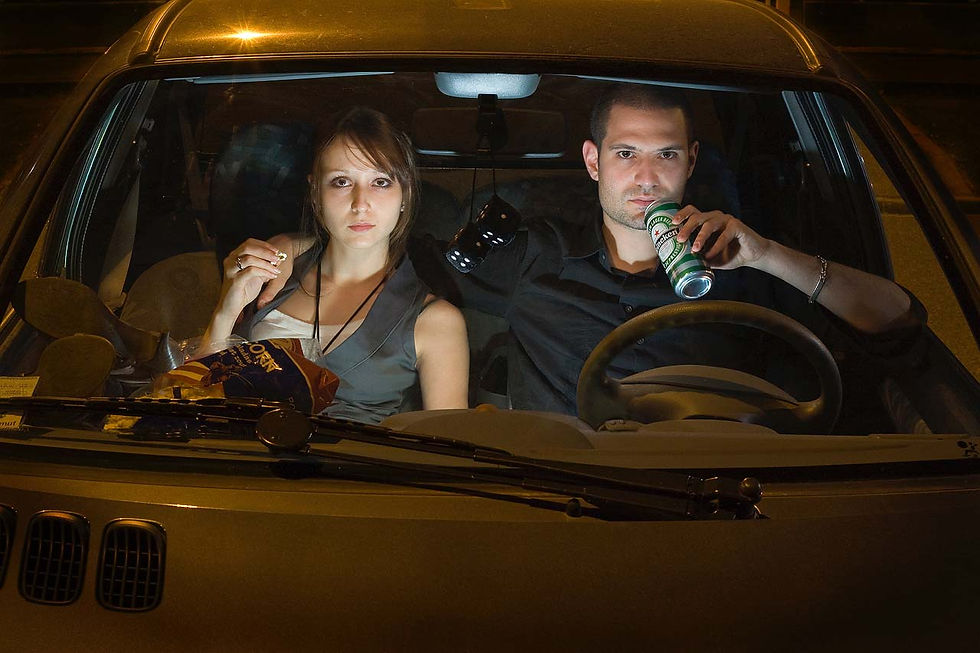A man holding a drink and a woman eating popcorns, sitting in a car, at night.