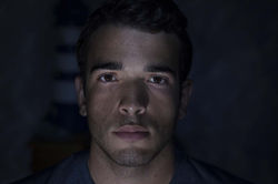 Face 9. Horizontal color close-up of a man looking directly at the camera, with low-key indoor night