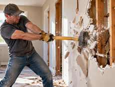Man in cap and gloves swings a sledgehammer at interior drywall during home demolition, with debris flying.