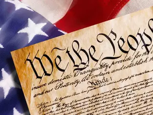 Close-up of the U.S. Constitution's "We the People" text on parchment, with the American flag's stars and stripes in the background. Patriotic mood.