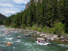 White raft with people navigating a rocky river through pine forest and cliffs under a partly cloudy sky