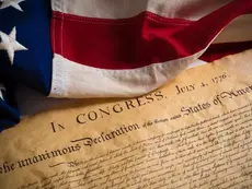 American flag draped over the Declaration of Independence document dated July 4, 1776. The flag's colors are vivid red, white, and blue.