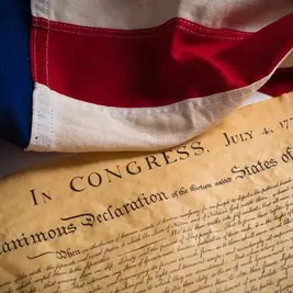 American flag draped over the Declaration of Independence document dated July 4, 1776. The flag's colors are vivid red, white, and blue.
