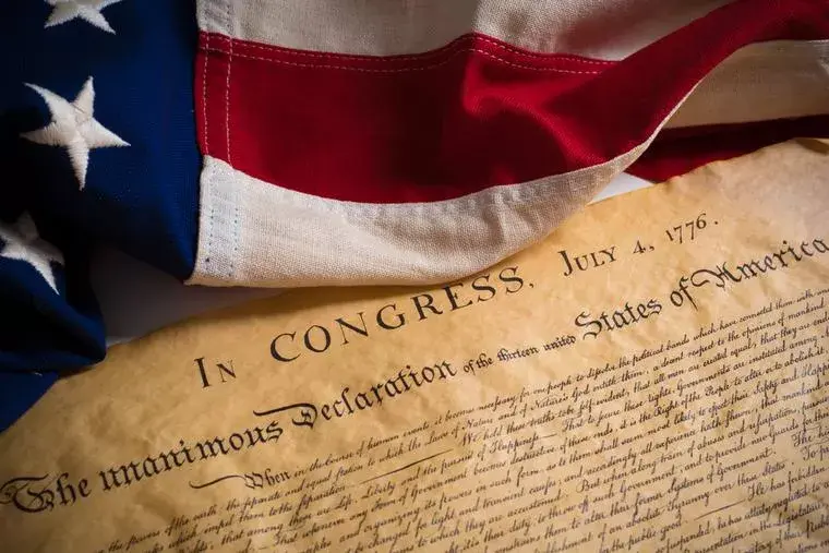 American flag draped over the Declaration of Independence document dated July 4, 1776. The flag's colors are vivid red, white, and blue.