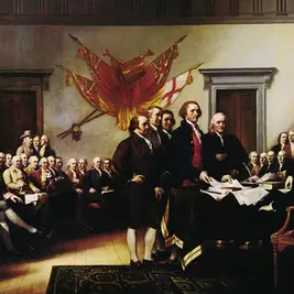 Delegates in a colonial chamber gather around a table signing documents, with red drapes and a patriotic banner on the wall.