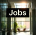 Black hanging sign reading Jobs in a bright glass office lobby with sunlight and blurred seating in the background.