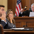 Shocked man and woman cover their mouths in a courtroom as a judge raises a gavel and reads a document, with a U.S. flag behind.