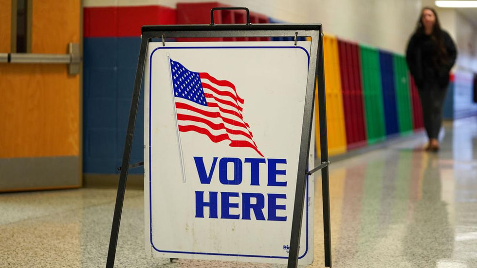 Vote Here sign with U.S. flag in a school hallway, rainbow wall panels behind it and a blurred woman walking in the background.