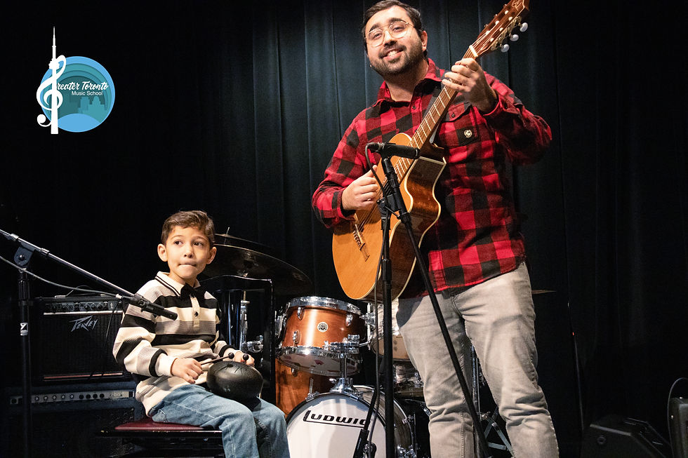 A man in red plaid plays guitar next to a boy holding a percussion instrument on stage. Drum set in background. Logo reads Greater Toronto Music School.