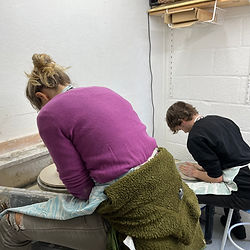 Back views of two people in a pottery lesson sat at potter's wheels throwing pots