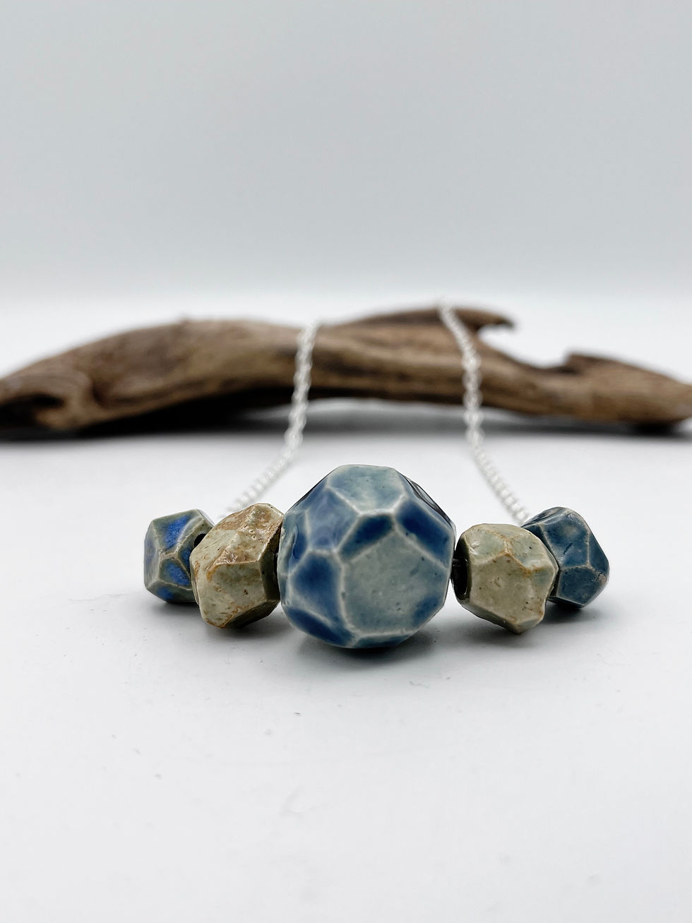 Thumbnail: Front view of a 5 faceted ceramic bead necklace resting on a piece of driftwood