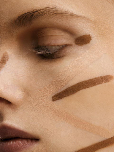 Close-up of a model's face with closed eyes, featuring various shades of foundation or contouring makeup swatched across the skin, showcasing a natural beauty aesthetic and makeup artistry.