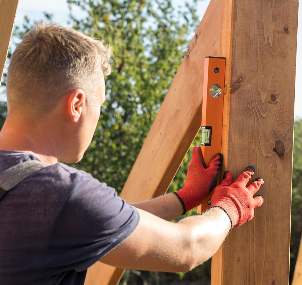 A person wearing red gloves uses an orange level on a wooden beam outdoors, under a sunny sky with trees in the background.