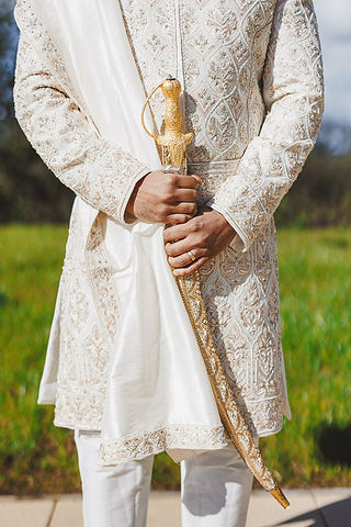 Man in embroidered cream suit holds a sword, wedding ceremony preparation.
