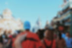 Crowd walking toward a fairytale castle down a themed street lined with decorated buildings under a clear blue sky. Festive atmosphere.