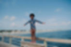 Child smiling with arms outstretched, stands on a railing in a floral swimsuit. Background of ocean, sky, and distant beachfront houses.