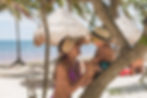 Woman in purple bikini and hat laughs with a toddler wearing a straw hat and sunglasses on a beach with palm trees and huts.