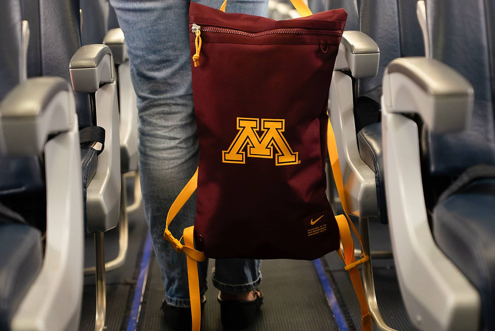Person standing in airplane aisle with maroon backpack showing yellow "M" logo. Blue jeans and seat rows visible. Air travel scene.