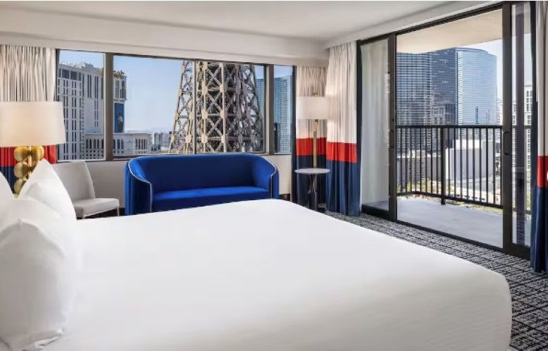 Hotel room with a white bed, blue couch, and city view. Large windows show a tower and buildings, creating a modern and bright atmosphere.