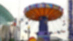Colorful swing ride at an amusement park with people enjoying the ride. The backdrop shows glass buildings and a clear sky.