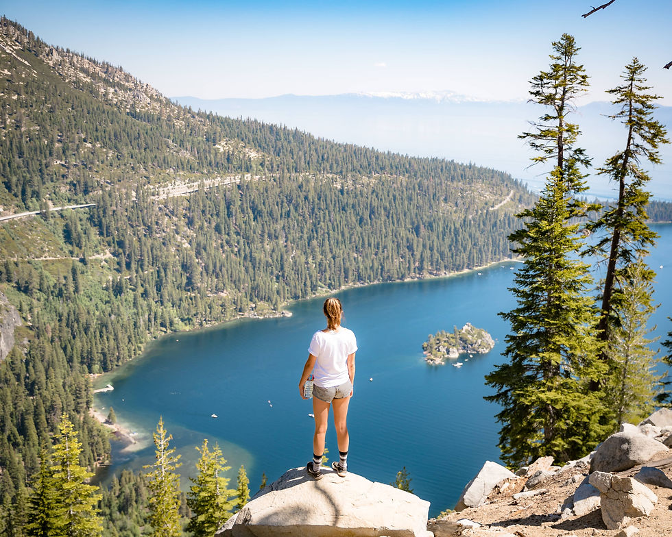 A person in shorts and a t-shirt stands on a rock overlooking a large blue lake surrounded by pine-covered mountains under a clear sky.