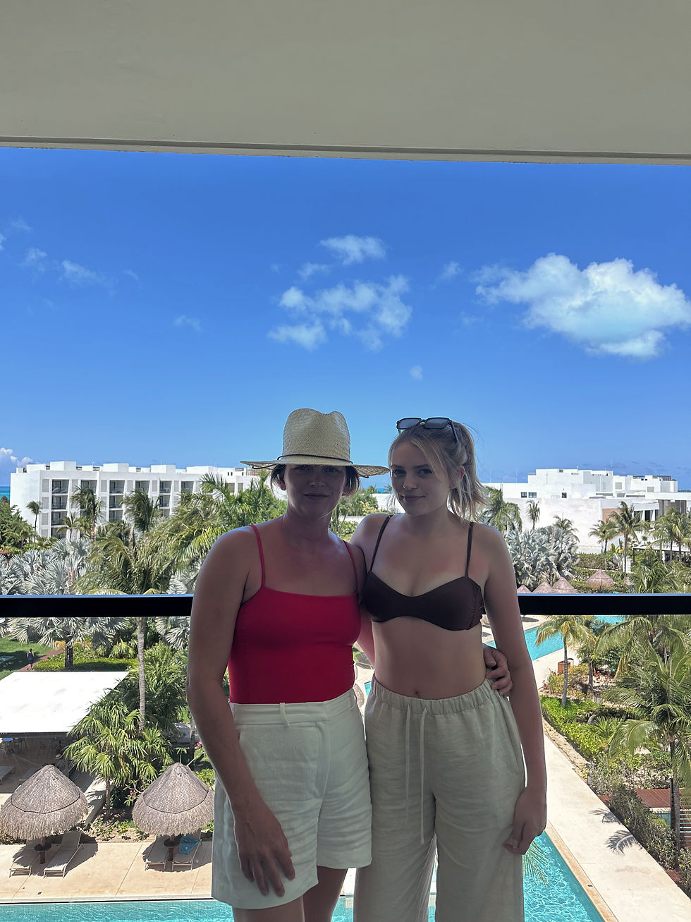 Two women on a balcony in Playa Mujeres wearing neutral vacation outfits—one in a red one-piece with linen shorts and straw hat, the other in relaxed beachwear—overlooking palm trees, pool, and ocean.