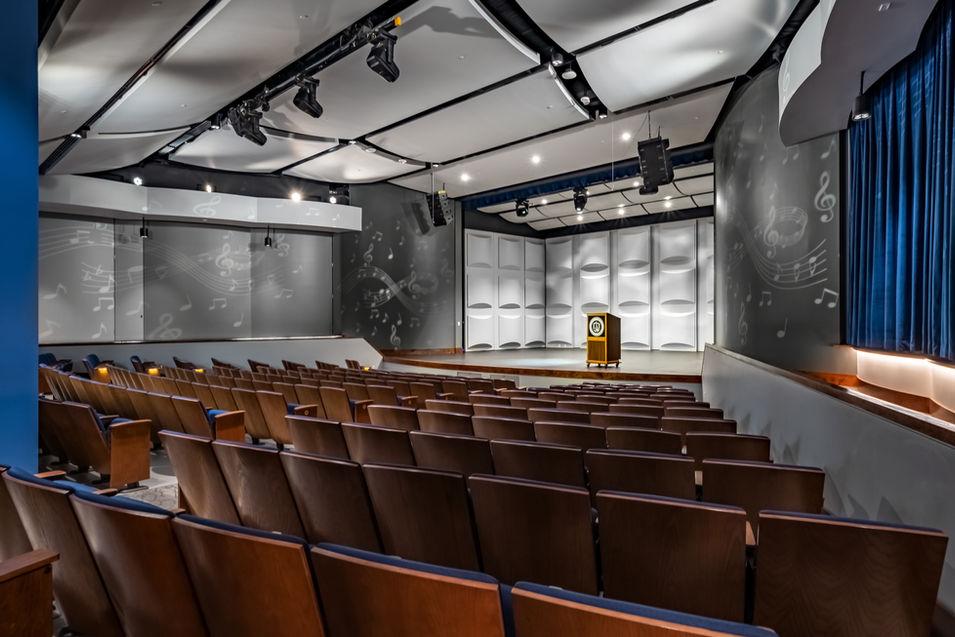 Empty auditorium with rows of chairs facing a stage with a podium , backdrop, and decorative acoustical panels.