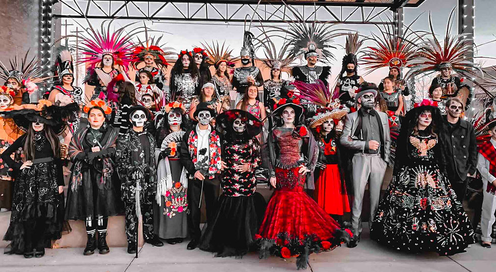 Group in colorful Día de los Muertos costumes and face paint, vibrant feathered headdresses, posing on a lit stage, festive mood.
