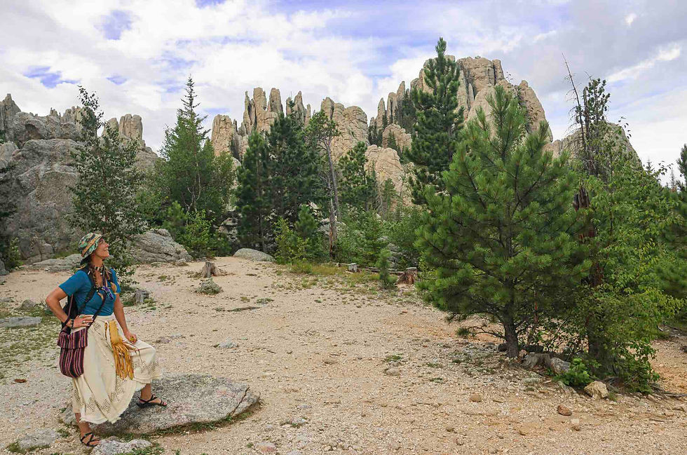 A woman in a blue shirt stands on a rock, looking at rock formations and pine trees under a cloudy sky. She wears a hat and skirt.