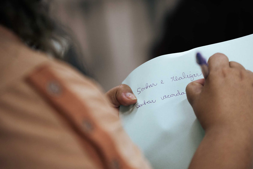 Fotografia colorida, em detalhe. Em desfoque, mão segura folha branca, a outra mão segura uma caneta. No papel sulfite, se-lê: sonhar e realizar. Sonhar acorda.