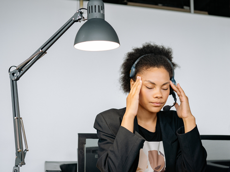 A black female business owner wearing a headset and a black blazer sits at a desk, pressing her fingers to her temples with her eyes closed, appearing stressed or dealing with a headache