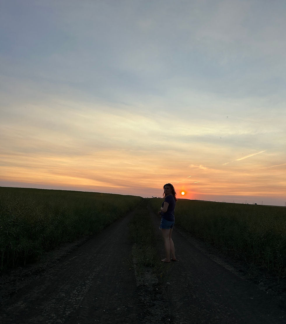 A woman stands barefoot on a prairie dirt road at dusk, holding her shoes in one hand, facing the glowing orange sunset. The wide sky and darkening fields frame her in silhouette, evoking solitude, longing, and quiet strength.