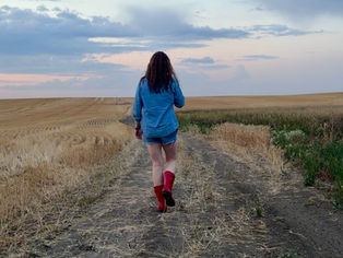 Woman in red rubber boots walking away down a prairie dirt road beneath a wide late-summer sky.