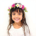 Smiling girl with long hair wears a colorful flower crown and white shirt against a plain white background. Cheerful and vibrant scene.