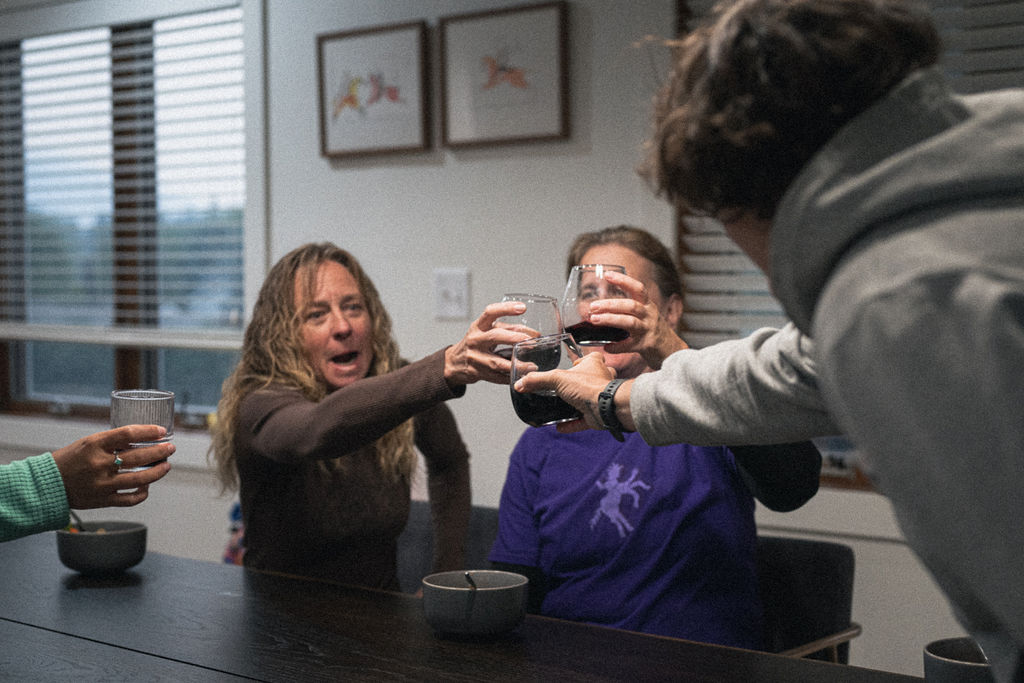Three women in casual clothes clinging wine glasses together in celebration in a softlit room