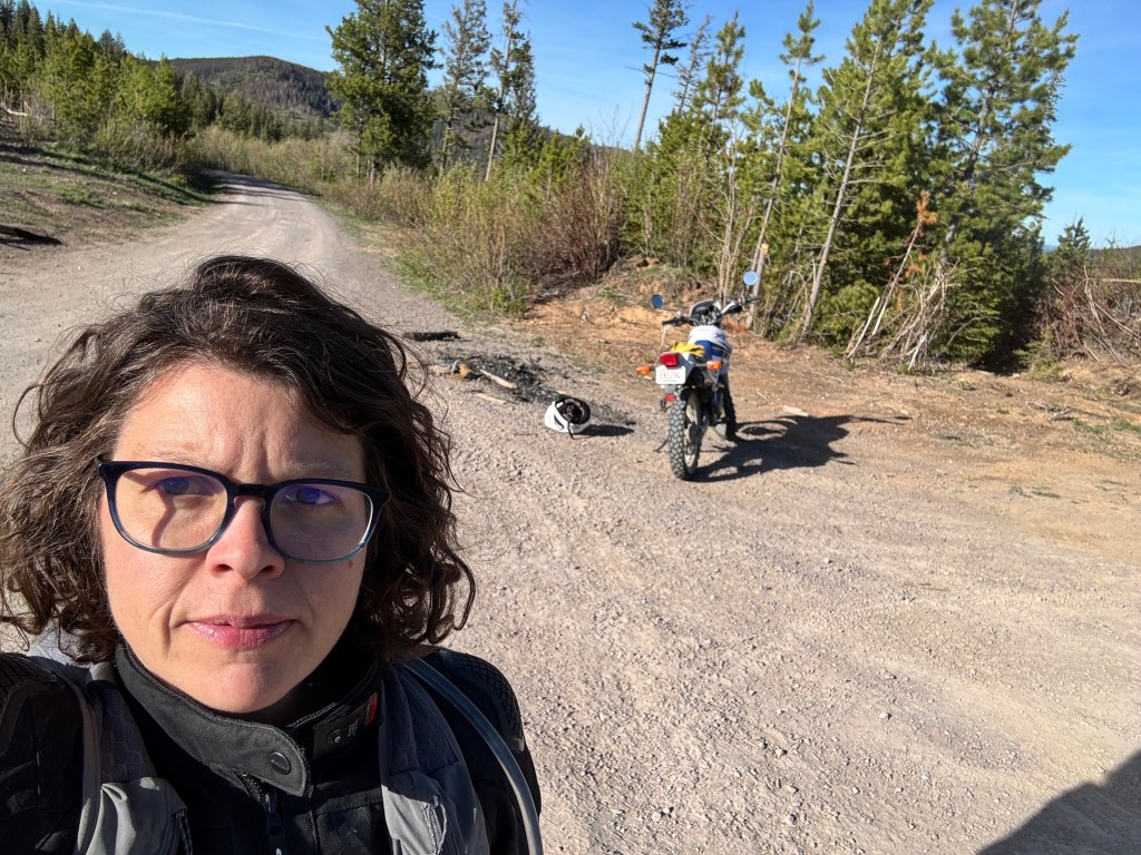 A person with curly hair and glasses wearing a motorcycle jacket stands on a dirt road, with a motorcycle parked nearby in a forested area.