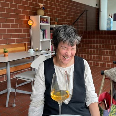 Jennifer Bartlett, a woman with short black and gray hair that falls into her face, is laughing. She's seated with a glass of wine in front of her, in a room with brick walls and bookshelves in the background.