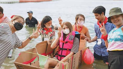 กลุ่มพนักงานกำลังสนุกสนานกับกิจกรรม Amazing Race โดยมีการต่อเรือจากกล่องกระดาษและทำกิจกรรมในน้ำร่วมกัน