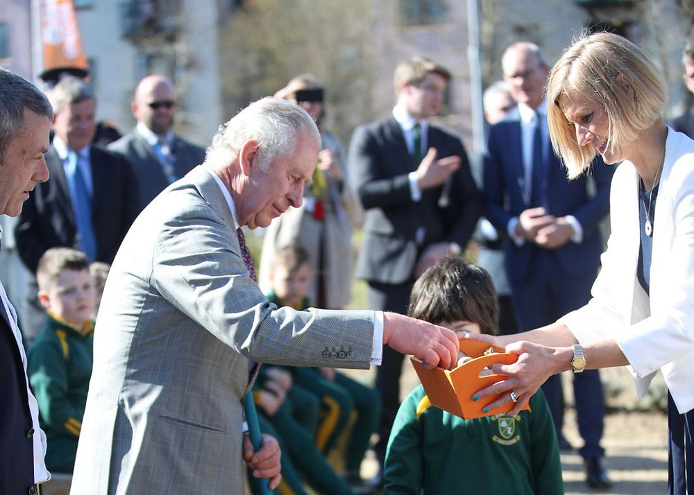 Prince Charles has right arm extended with right hand in a try as if reaching for something. Minister Hackett is holding the tray while he does this. They are out of doors and a number of adults and children are looking on.