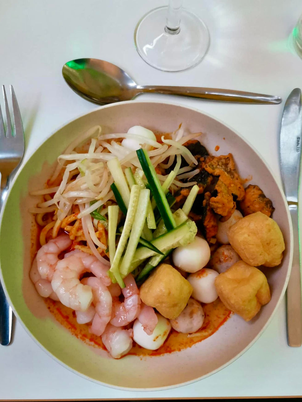 "A bowl of vibrant Singaporean laksa featuring shrimp, bean sprouts, cucumber slices, tofu cubes, quail eggs, and meat in red sauce, served on a white table with cutlery, a wine glass, and a glass of water."
