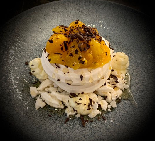 A plated pavlova-style dessert featuring a crisp meringue shell filled with whipped cream, topped with passionfruit and dark chocolate shavings. Surrounding the centerpiece are broken meringue pieces and dollops of cream, artfully arranged on a textured dark gray plate. The vibrant yellow fruit contrasts with the neutral tones, creating an elegant and visually striking presentation.