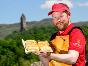 A man in a red Bayne's uniform with a tray of sausage rolls