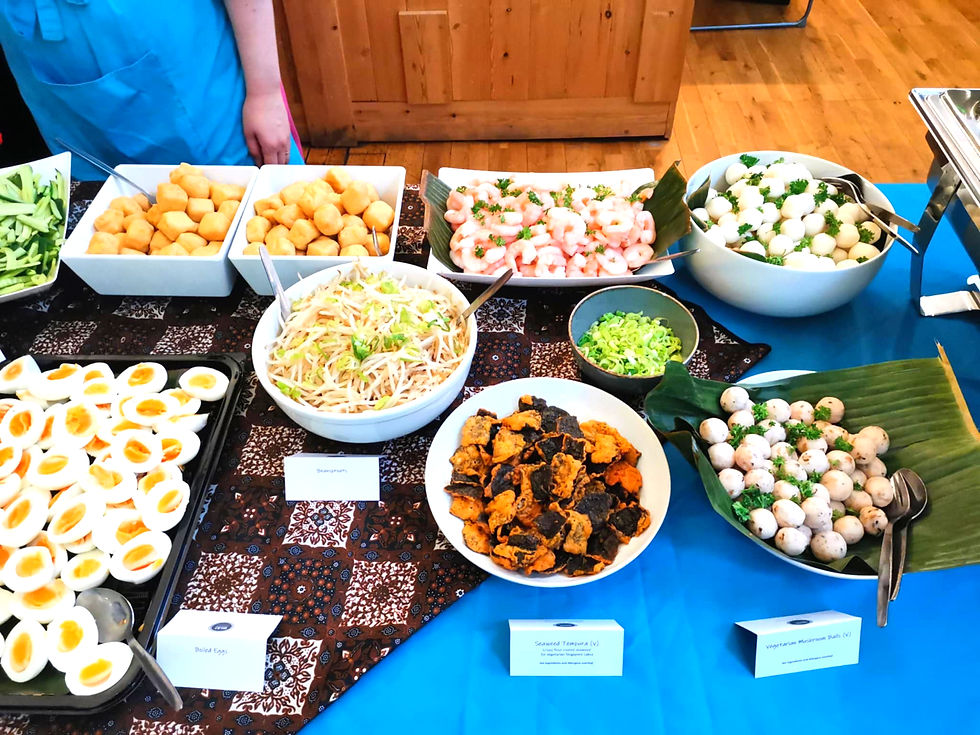 "Buffet table with trays of sliced cucumbers, fried tofu cubes, shrimp, fish balls, halved boiled eggs, bean sprouts, tofu skin, and chopped green onions, set on a blue and brown patterned cloth with labeled dishes and two apron-clad hosts behind."