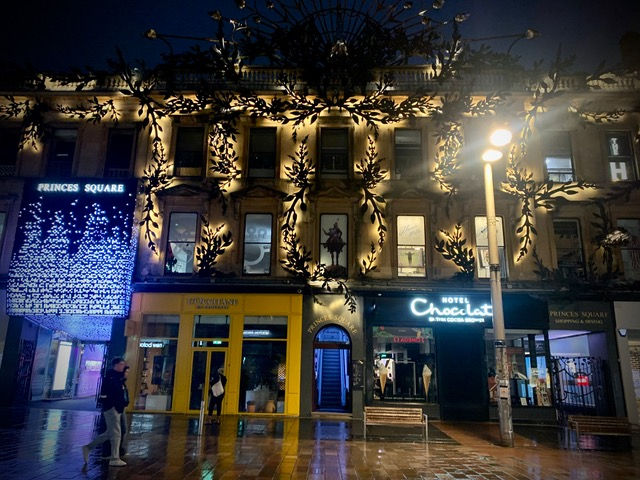  Nighttime view of a stylish urban Buchanan street in front of Princes Square, featuring ornate illuminated leaf and branch designs on the building facade. The ground floor showcases brightly lit signs for Hotel Chocolat and other shops. A cascading light display with the text "PRINCES SQUARE" adds drama to the scene. Reflections shimmer on the wet pavement as a few pedestrians pass by, capturing the vibrant, artistic energy of the city.
