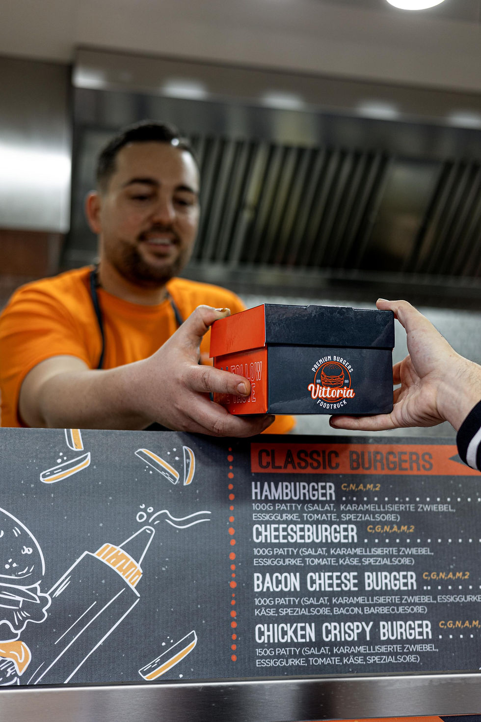 Mitarbeiter vom Foodtruck Vittoria übergibt Burger-Box an Kunden; im Vordergrund Menütafel mit Classic Burgers.